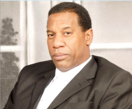 A man in a dark suit and white shirt sits indoors, looking at the camera with a neutral expression.