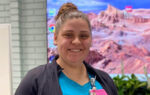 Smiling person wearing a blue shirt, black jacket, and name badge, standing indoors with a colorful desert landscape image displayed in the background.