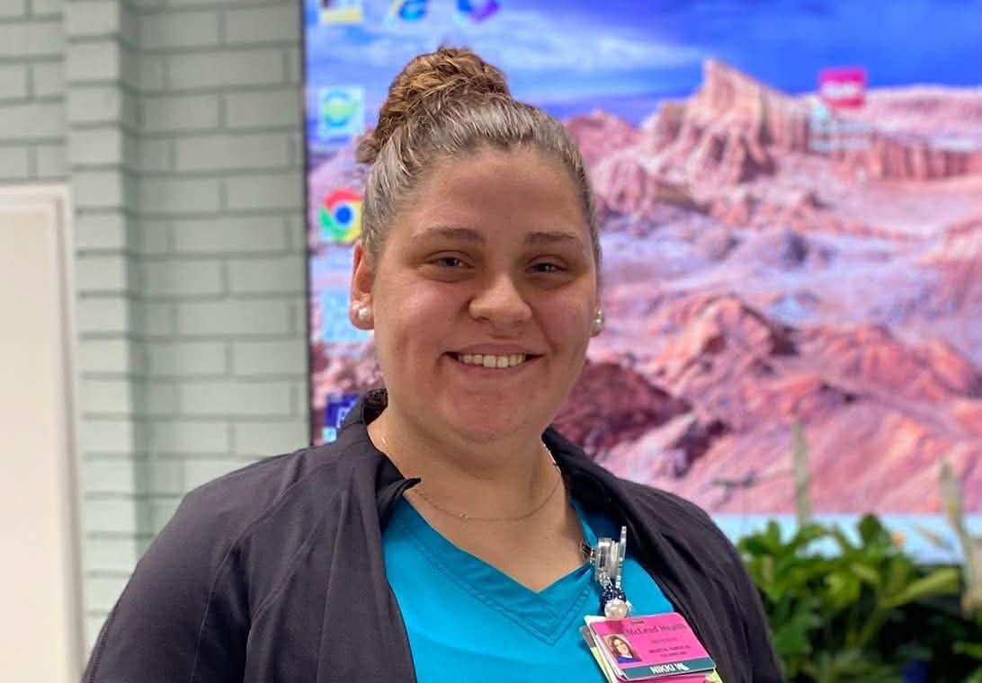 Smiling person wearing a blue shirt, black jacket, and name badge, standing indoors with a colorful desert landscape image displayed in the background.