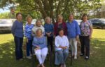 A group of ten women pose outdoors under a tree, with two seated in front and eight standing behind, smiling at the camera.
