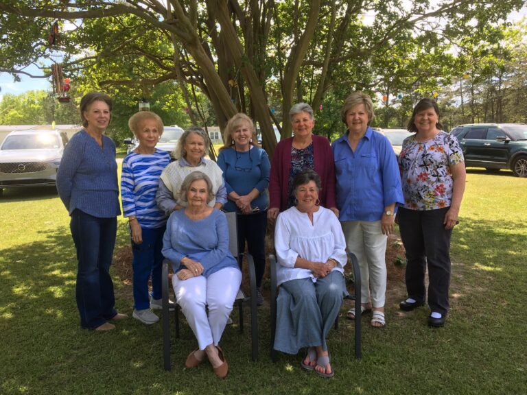 A group of ten women pose outdoors under a tree, with two seated in front and eight standing behind, smiling at the camera.