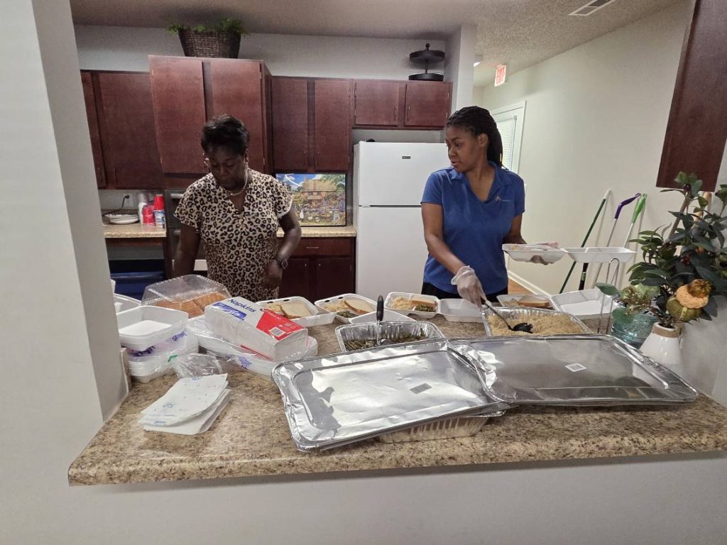Two women prepare and package food in a kitchen, with trays, containers, and utensils spread out on the counter.