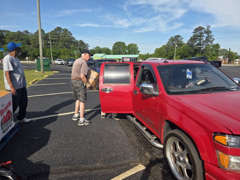 A man loads a cardboard box into a red SUV parked in a sunny parking lot with trees and cars in the background.