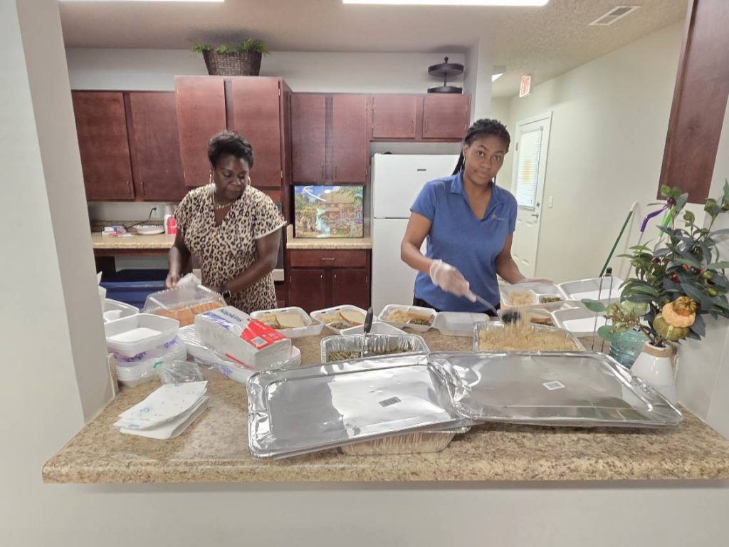 Two women in a kitchen packing food into containers, surrounded by trays, boxes, and kitchen supplies on a countertop.