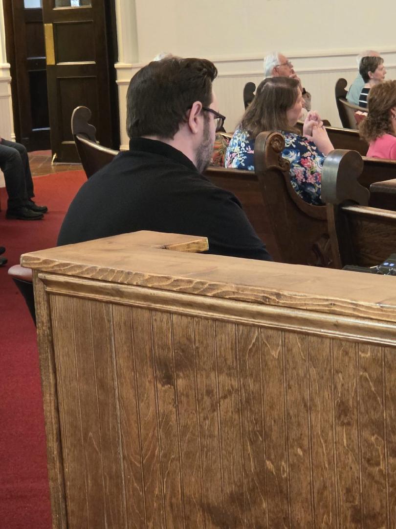 People sitting in wooden pews inside a church, some engaged in conversation, on a red carpeted floor.
