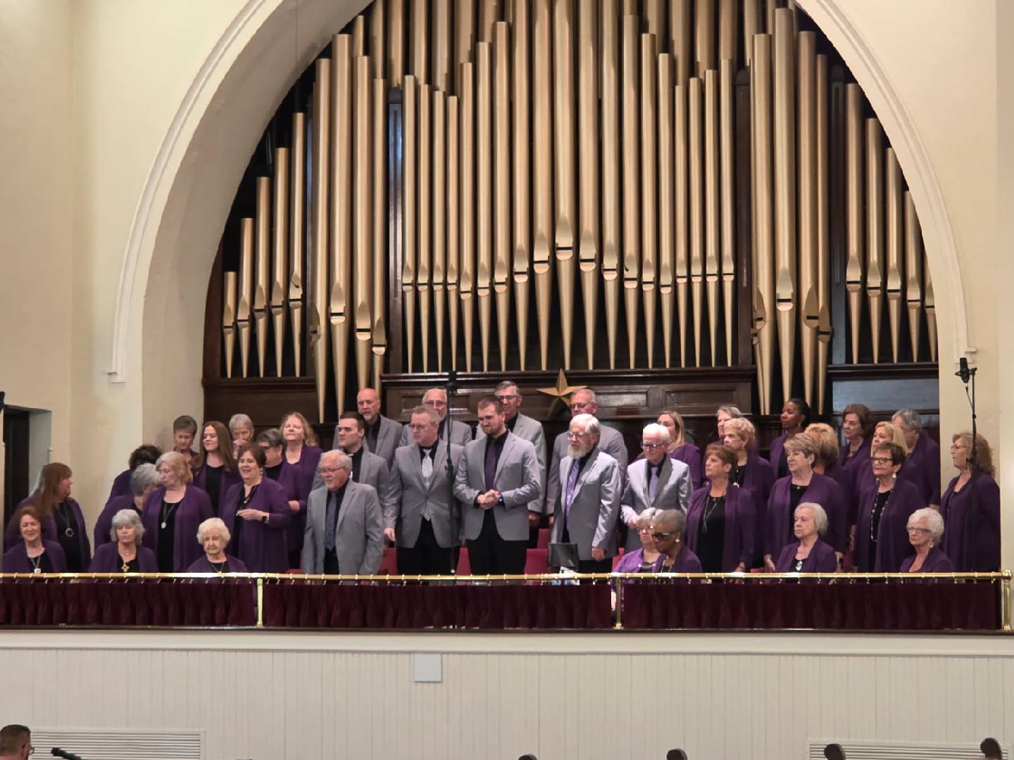A choir group stands on a balcony in front of a large pipe organ inside a church, dressed in purple and gray attire.