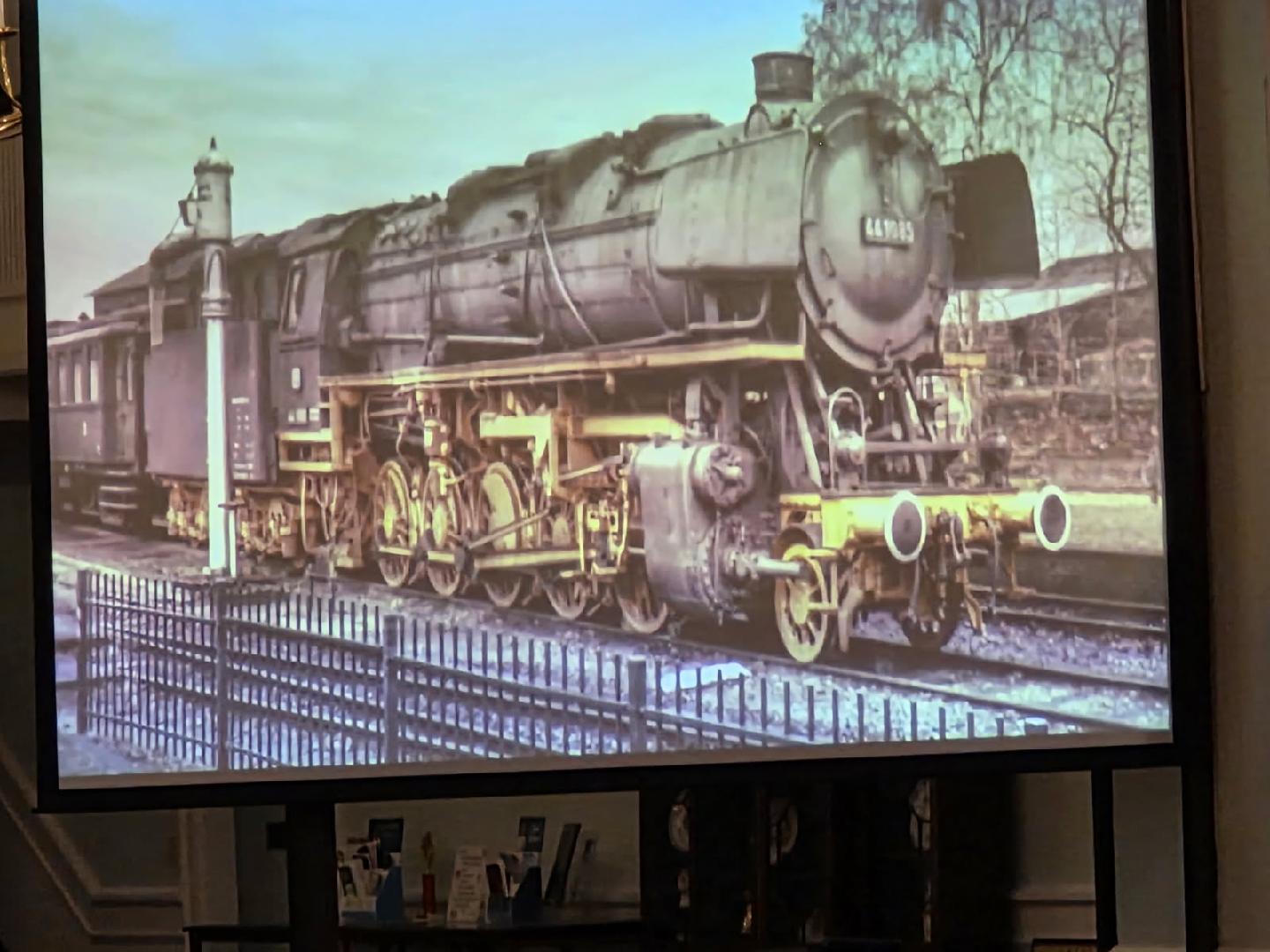 A vintage steam locomotive on display, viewed behind a railing, with trees and a building visible in the background.