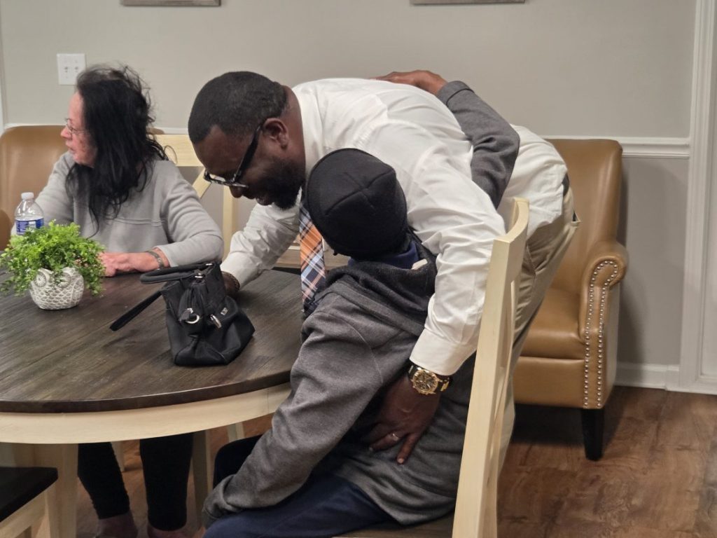 A man in a white shirt hugs a seated person, while a woman sits at a table with a plant and a purse.