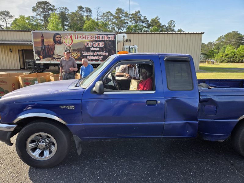 A blue pickup truck with a dented door, a person in the driver&rsquo;s seat, and people standing nearby in an outdoor setting with trees and a building.