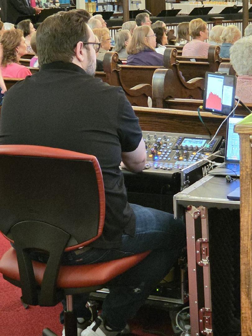 A person sits at a sound mixing console with multiple screens in a church or auditorium, managing audio during an event.