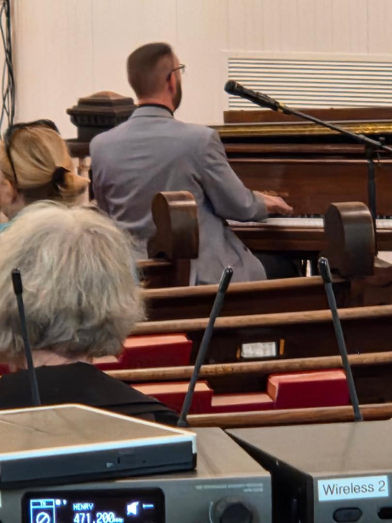 People seated in church pews, with a man in a gray suit playing piano at the front, microphones visible in the foreground.