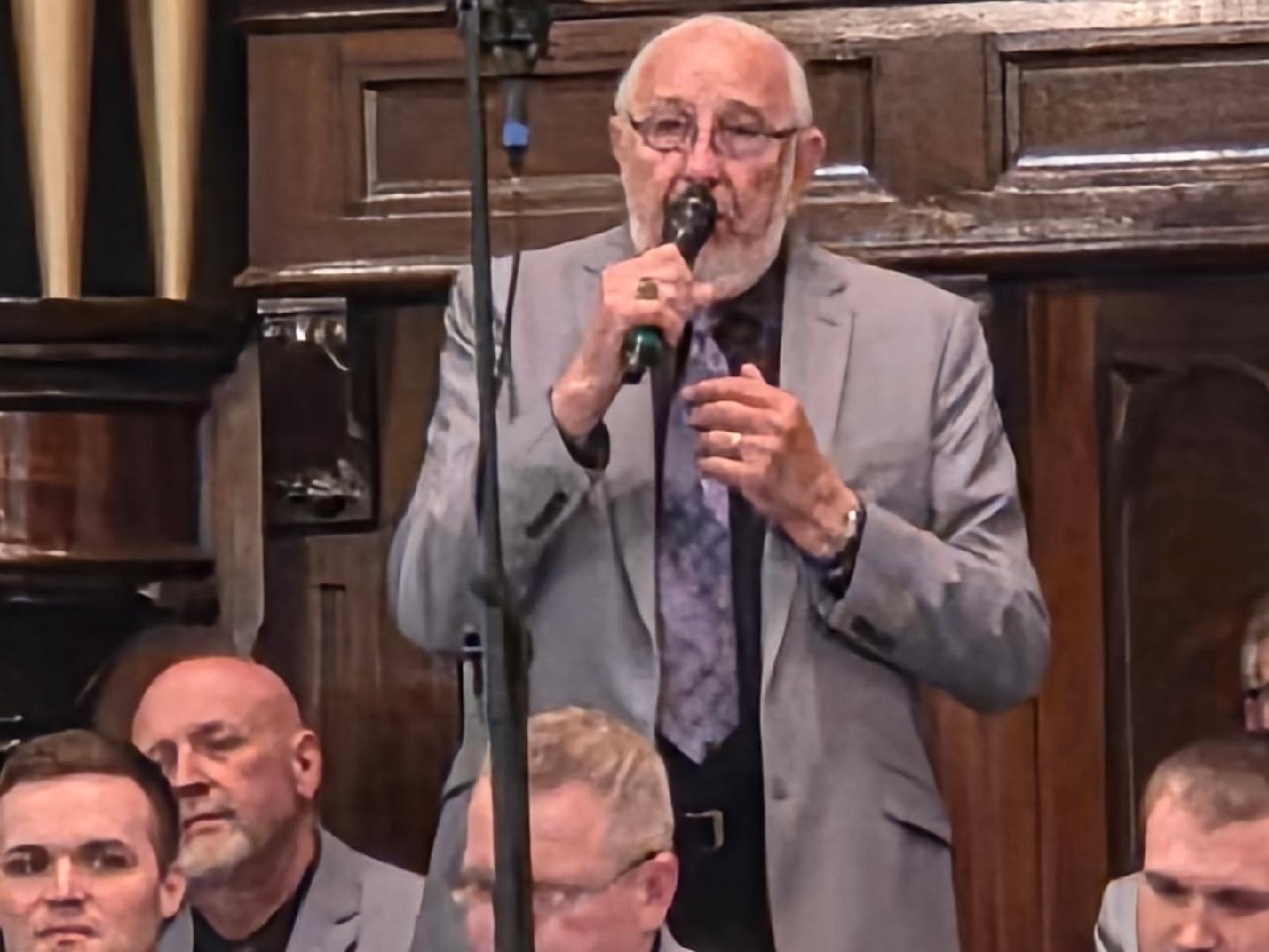 older man in a gray suit and glasses sings into a microphone indoors, with two other men seated nearby in similar attire.