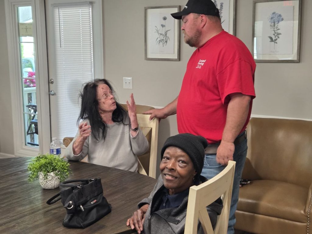 Two women sit at a table talking with a man in a red shirt and black cap, inside a well-lit room with framed flower prints.