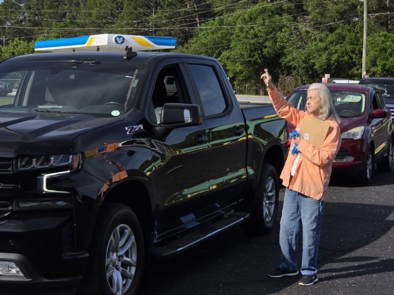 elderly person in an orange shirt stands beside a black pickup truck, pointing at something while outdoors. Trees are visible in the background.