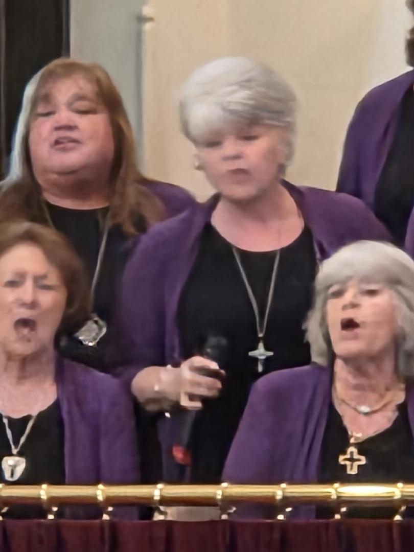 Four older women in matching purple shawls and black tops singing in a choir, each wearing necklaces, one holding a microphone.