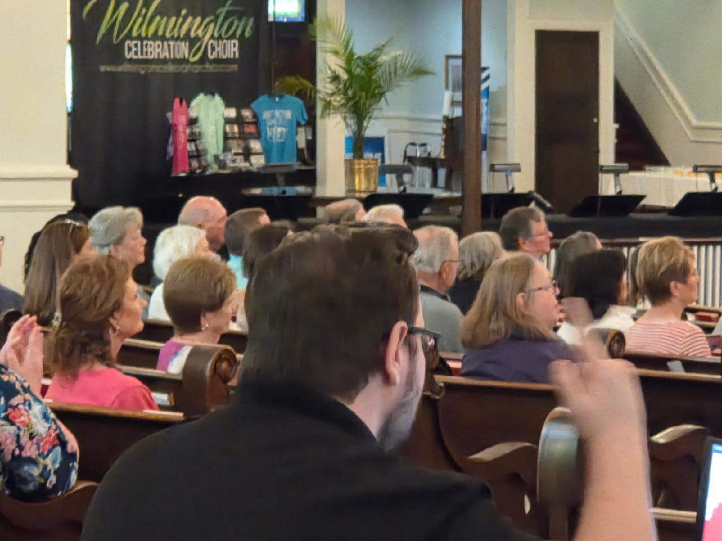 A group of people seated in a church or auditorium, listening to a speaker. There are shirts and signs displayed on a stage in the background.