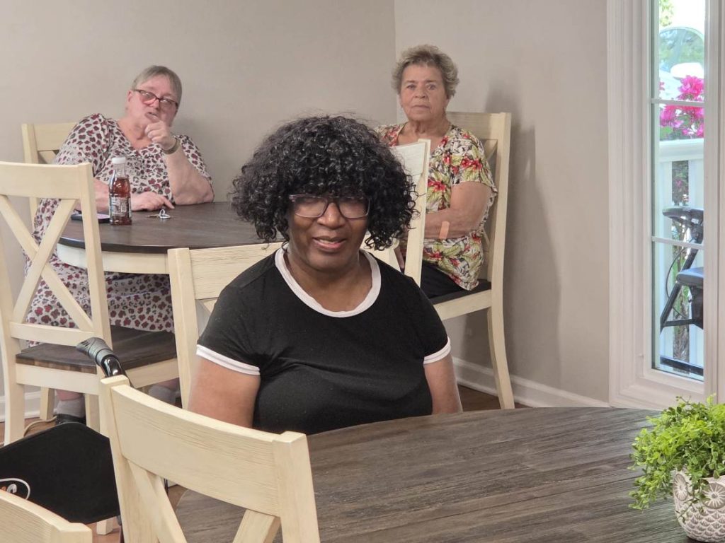 Three women sit at wooden tables in a bright room, two in the background and one smiling in the foreground.