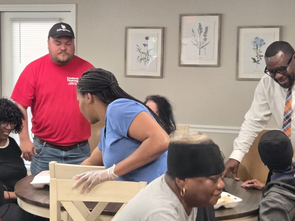 A group of people gathered around a table indoors, some seated and some standing, sharing food and interacting in a casual, friendly setting.