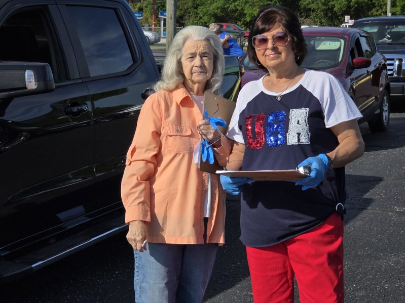 Two women standing outdoors by a vehicle, one holding a clipboard and both wearing casual clothes. The woman on the right has blue gloves, and it is sunny outside.