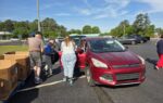 People loading items into a red car in a parking lot on a sunny day, with trees and other vehicles in the background.