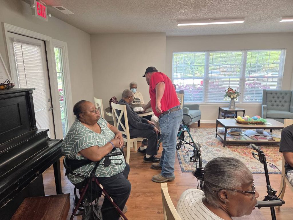 Several elderly people and a man in a red shirt gather in a bright common room, some using walkers, with a piano and large windows in the background.