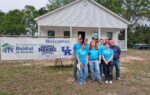 A group of people in blue shirts stand in front of a white house and a Habitat for Humanity welcome banner.