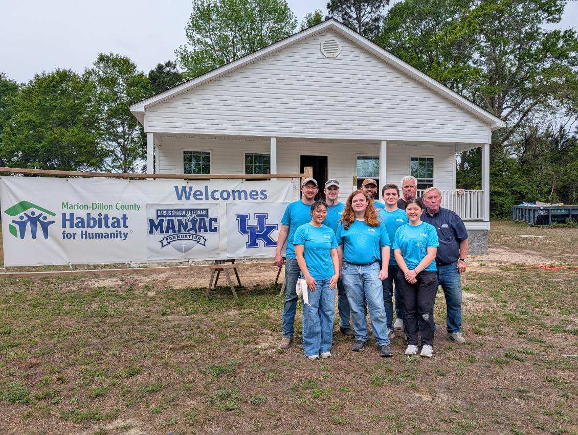 A group of people in blue shirts stand in front of a white house and a Habitat for Humanity welcome banner.