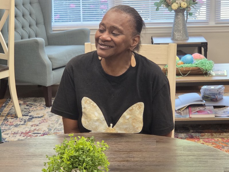 Smiling woman wearing a black shirt with a large yellow butterfly design, sitting at a table with a small green plant in a cozy room.