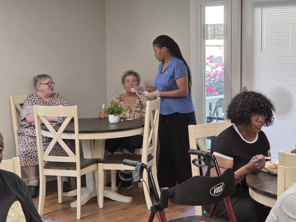 A group of elderly women sitting at tables in a cozy dining area, one woman serving food, another eating, and a walker in the foreground.