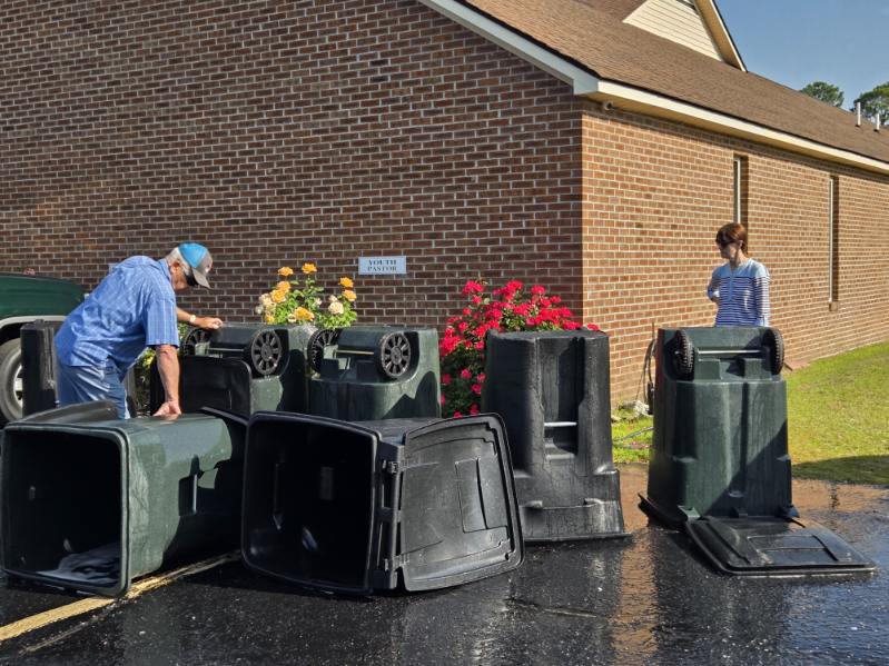 Two people stand outside by several overturned trash bins near a brick building with flowers and bushes visible on a sunny day.