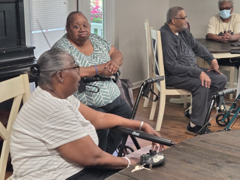 Four elderly people sit around a table in a room, some using walkers, with a relaxed and casual atmosphere.