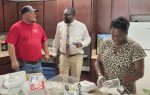 Three people in a kitchen prepare food; one woman is working with gloves, while two men are talking nearby.