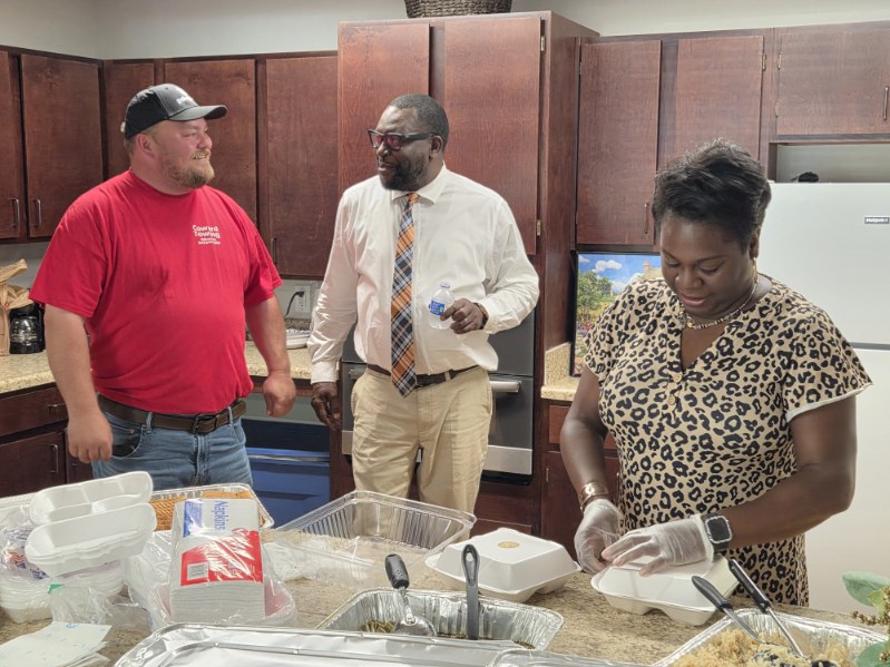 Three people in a kitchen prepare food; one woman is working with gloves, while two men are talking nearby.