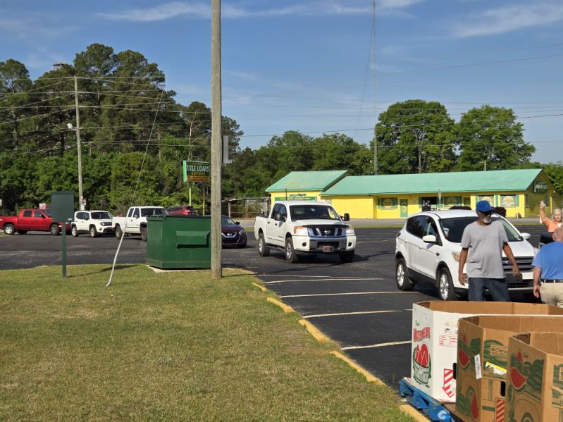 Cars lined up in a parking lot as a man in a cap walks near boxes, with a green and yellow building in the background.