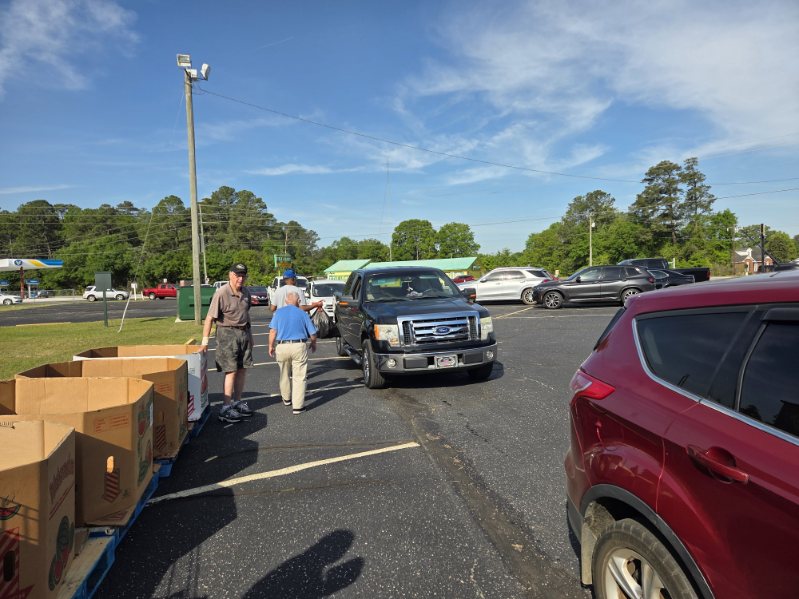 People standing and walking in a parking lot with several cars parked, under a blue sky with scattered clouds, and trees in the background.