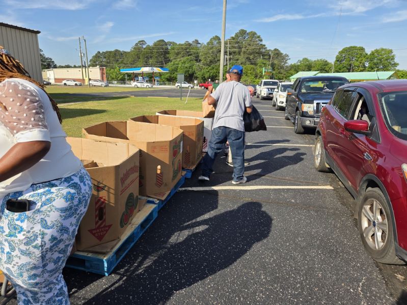 People distribute items from large cardboard boxes to cars in a parking lot during a drive-through event on a sunny day.