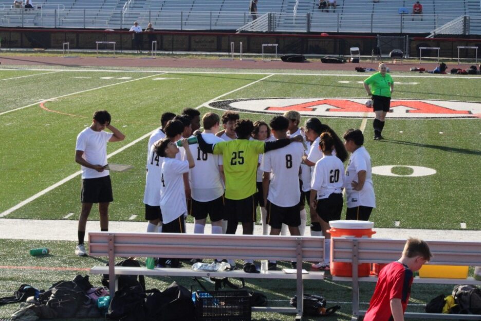 A soccer team in white uniforms huddles on the sideline of a sports field, listening to their coach. Bleachers and a referee are visible in the background.