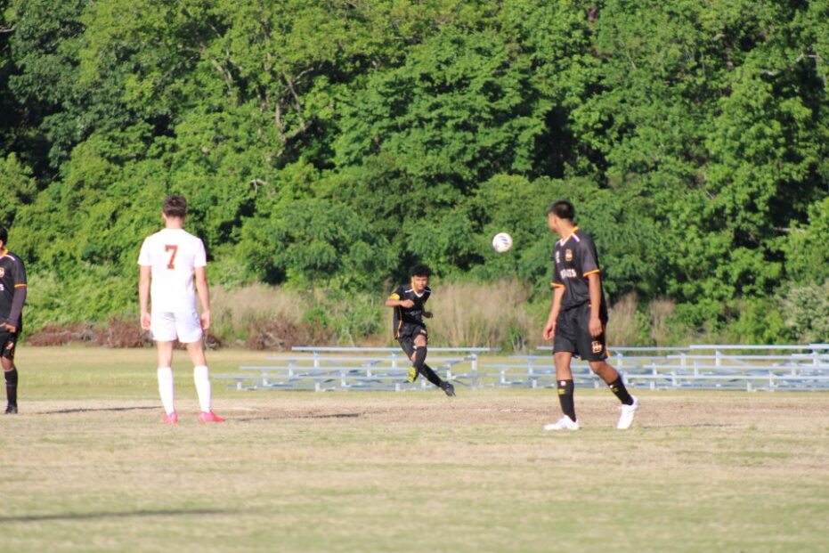 Soccer players compete on a grassy field; one player in black uniform kicks the ball, while others observe. Trees and empty bleachers are visible in the background.