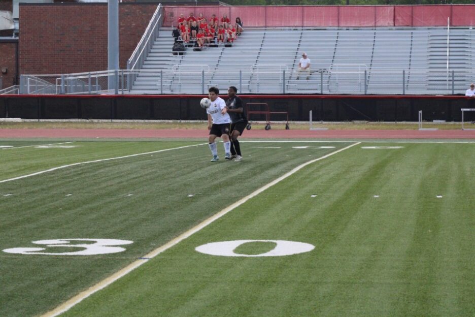 Two soccer players running on a field near the sideline, with empty bleachers and a few spectators in the background.
