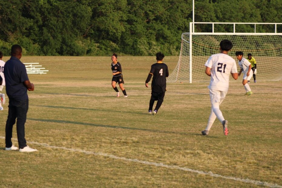 Soccer players in black and white uniforms play on a grassy field near a goal, with a coach watching from the sideline and trees in the background.