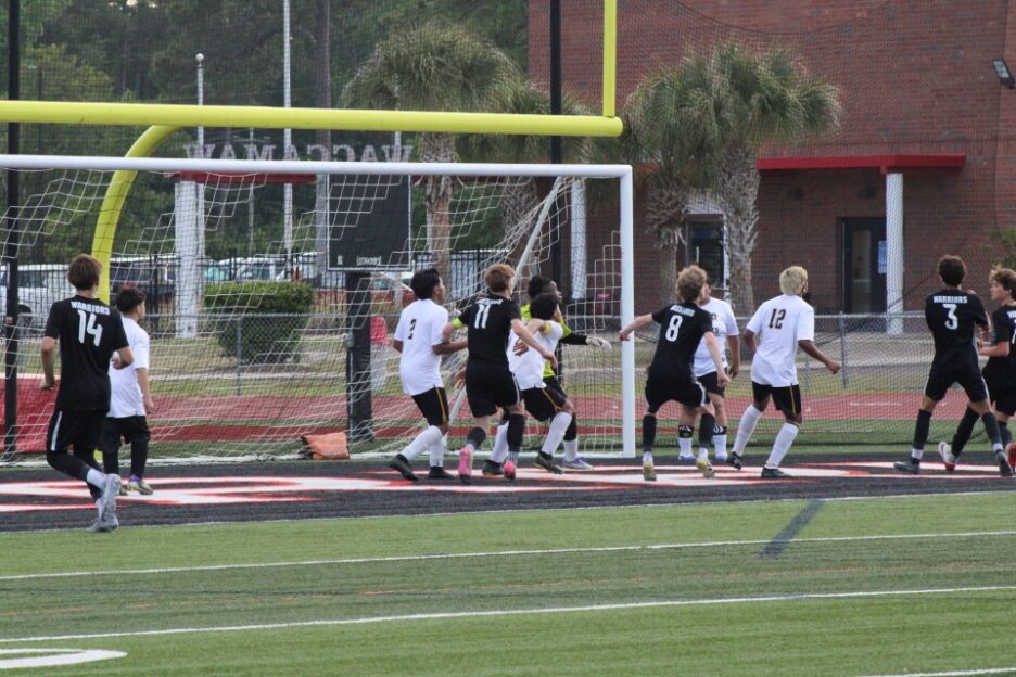 Soccer players in black and white uniforms gather near the goal during a match on an outdoor field.