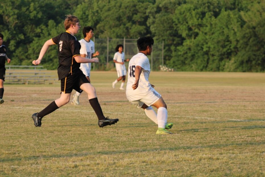 Two soccer players, one in black and one in white, compete for the ball on a grassy field with trees in the background.