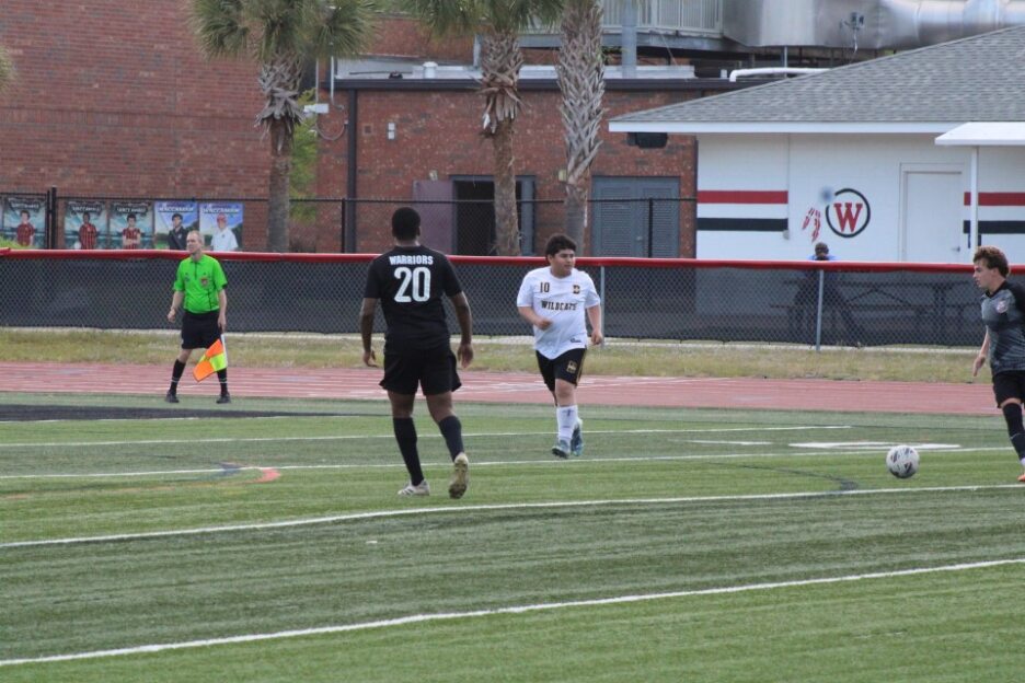 Two soccer players and a referee on a field, with a building and palm trees in the background.