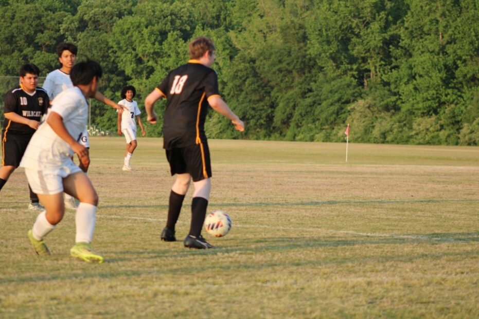 A group of soccer players compete on a grassy field, with trees in the background and a corner flag visible.