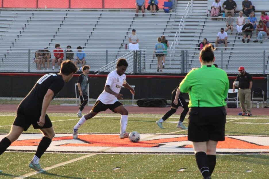 Soccer players competing for the ball on a field, with a referee observing and spectators seated in the background on stadium bleachers.