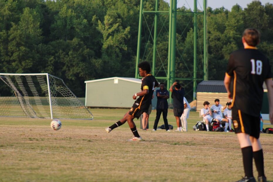 A soccer player in a black uniform kicks a ball on a field, with teammates, opponents, and a goal visible in the background.