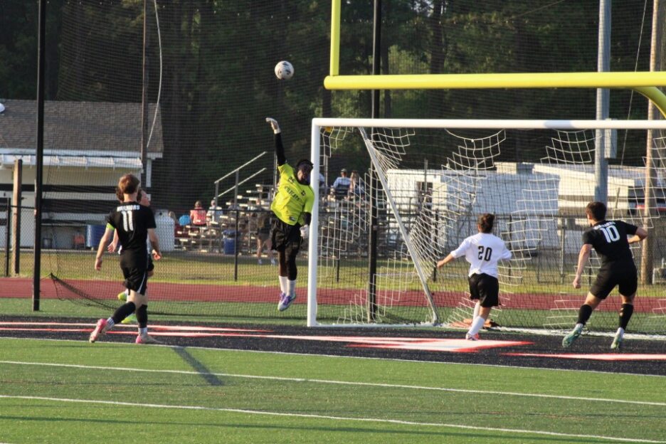 Soccer goalkeeper jumps to block a shot as players watch, near the goalpost on an outdoor field during a match.