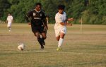 Two soccer players, one in black and one in white, chase the ball on a grassy field with trees in the background.