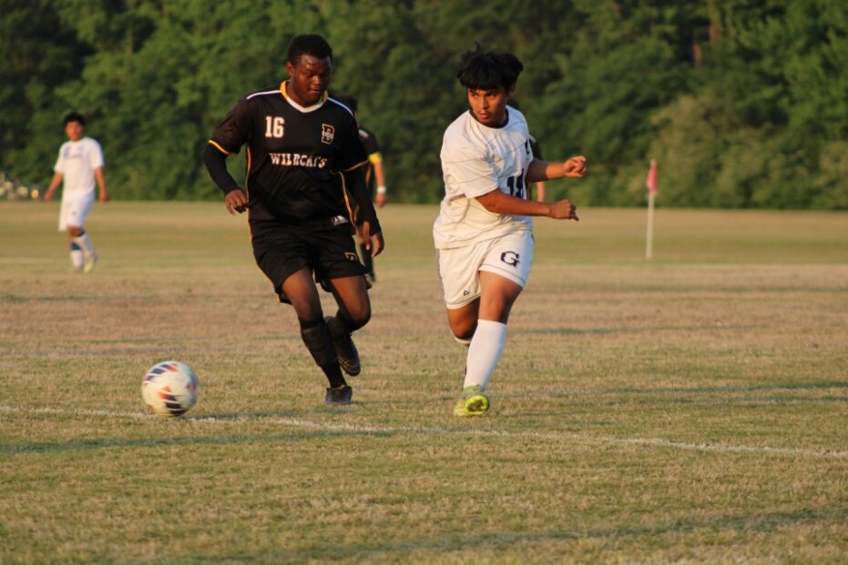 Two soccer players, one in black and one in white, chase the ball on a grassy field with trees in the background.