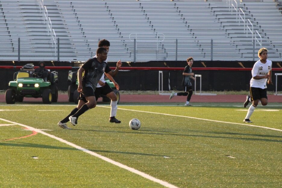 Soccer player dribbling the ball on a field during a game, with empty bleachers and other players in the background.